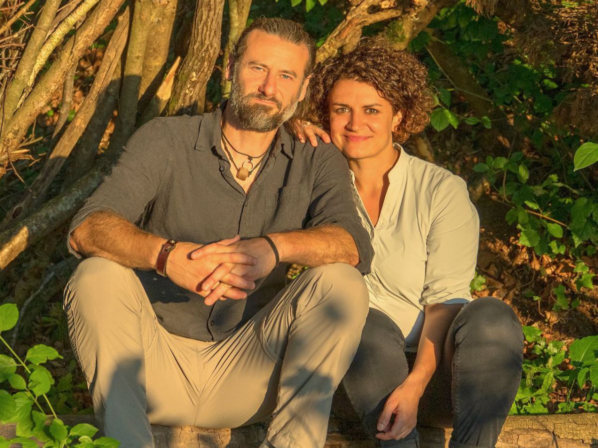 Ein Paar sitzt in der Abendsonne auf einem niedrigen Randstein im Grünen. Der Mann hat die Haare zusammengebunden und einen Bart, die Frau kinnlange lockige Haare. Sie lächeln in die Kamera.