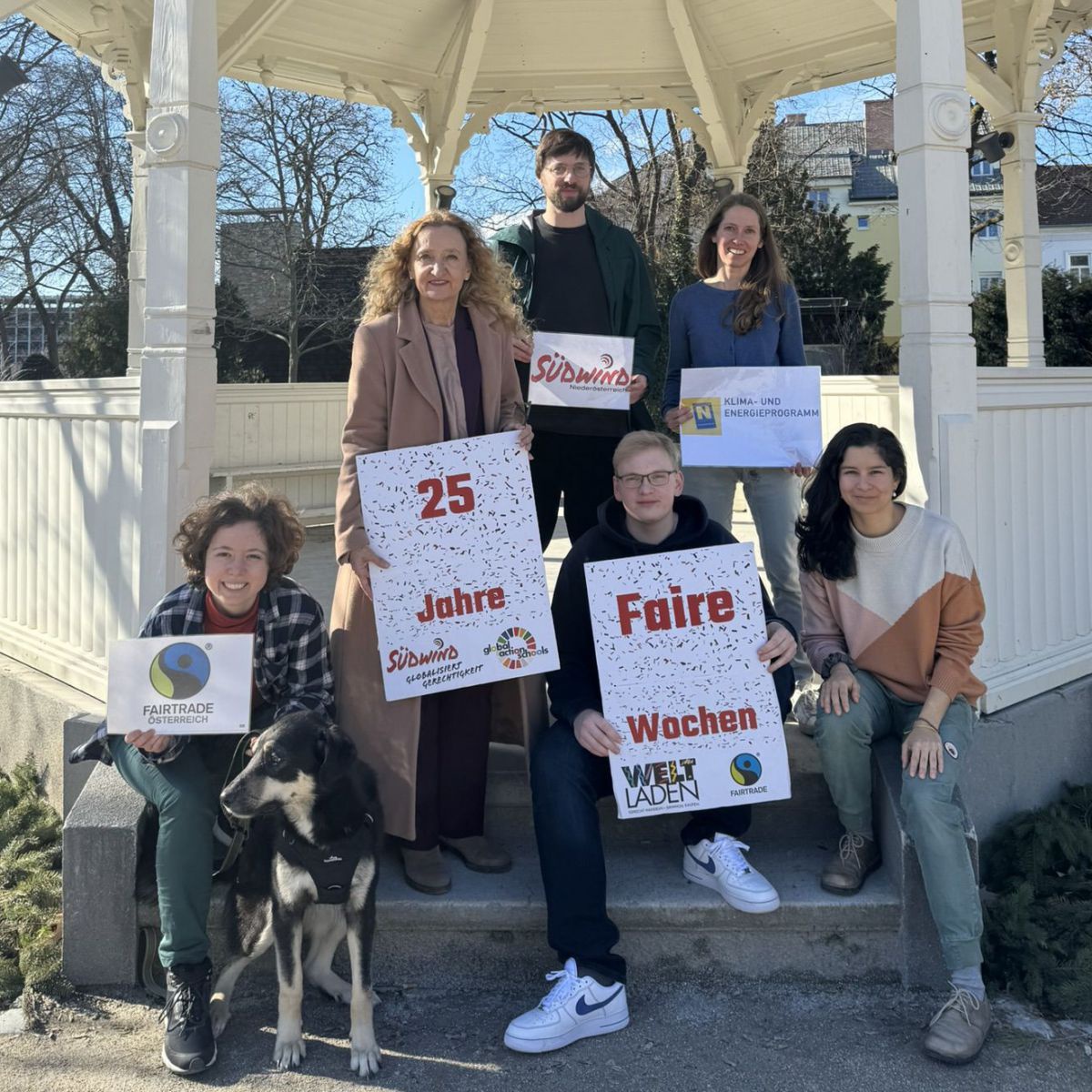 Unter einem weiß gestrichenen Holzpavillion ein einem Park posieren 4 Frauen, zwei Männer und ein Hund für das Foto. Einige zeigen Poster ins Bild, auf denen steht: 