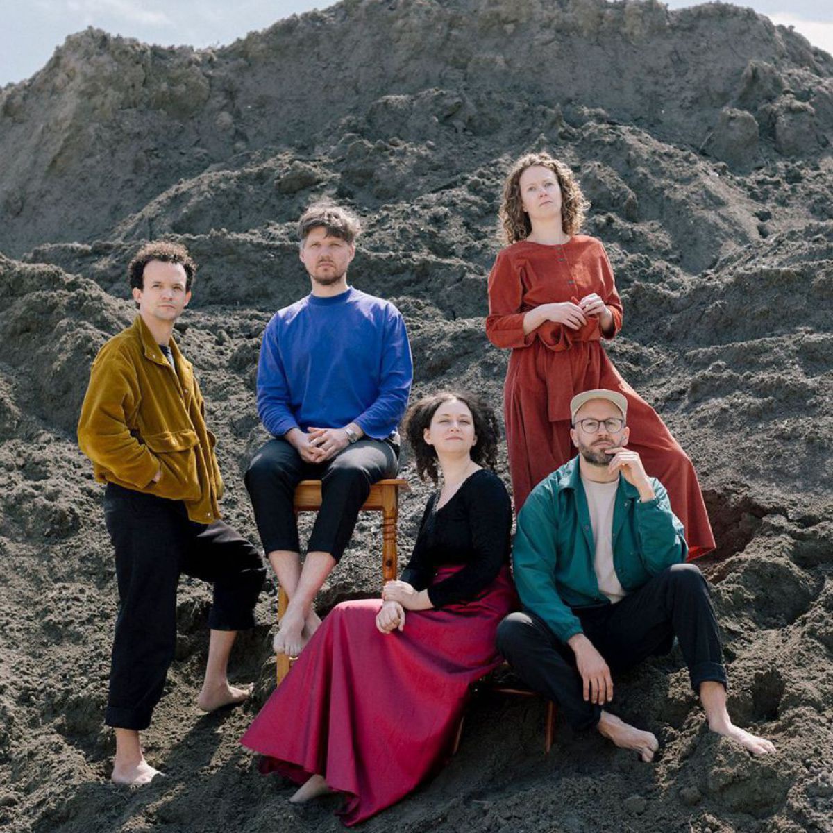 Die 5 Musiker*innen vom Ensemble SOIL posieren barfuß in bunten Gewändern am Hang eines riesigen Haufens dunkler Erde.