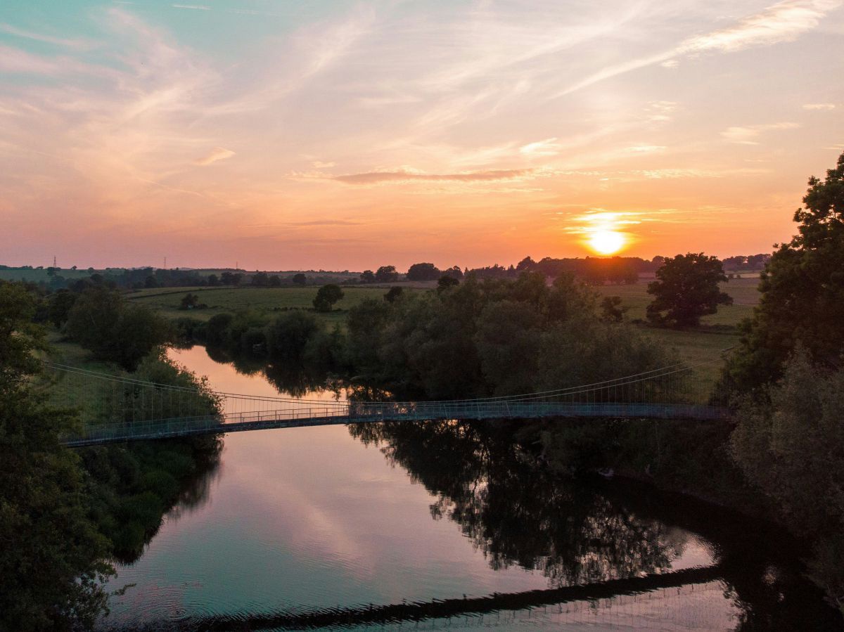 Sonnenuntergang über einer Landschaft, durch die ein breiter Fluss fließt. Eine fein gebaute Hängebrücke überspannt das Gewässer, welches an beiden Ufern dicht bewachsen ist.