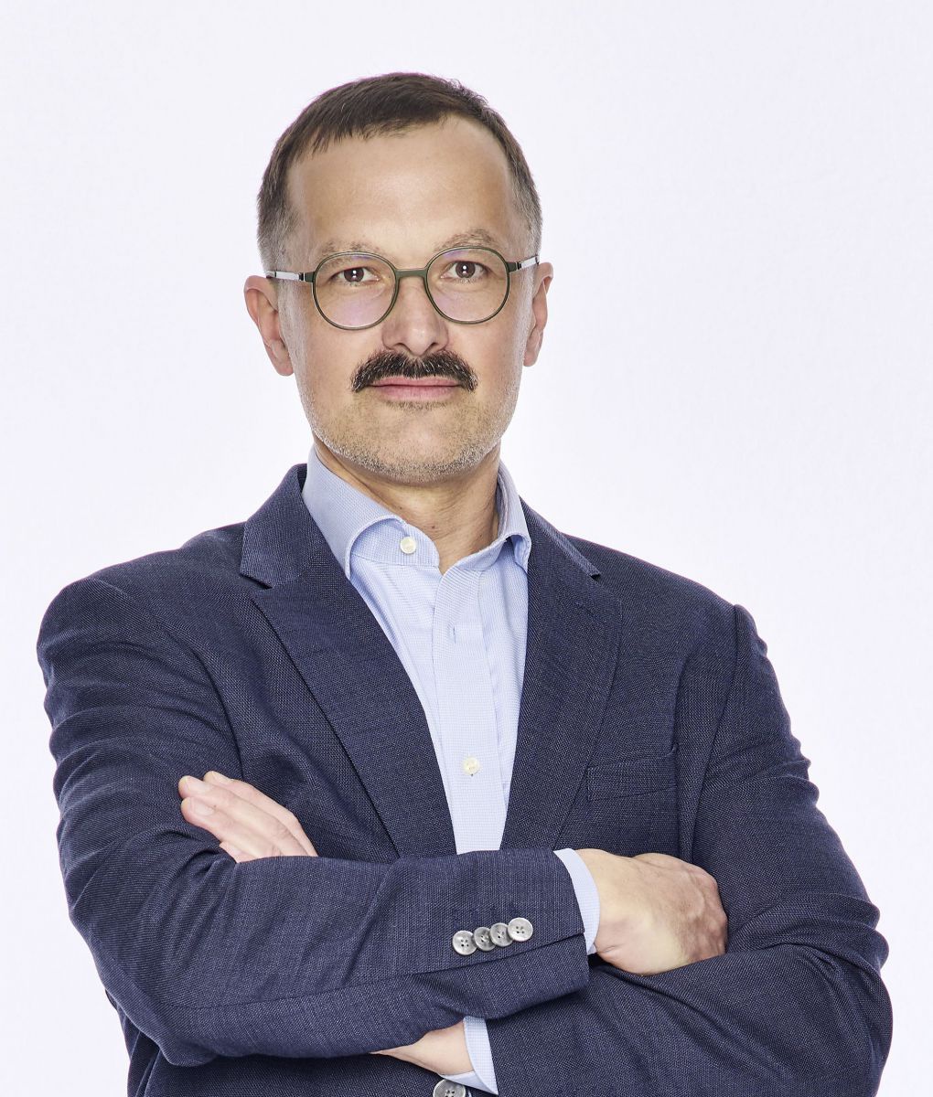 Mann mit kurzem, dunklem Haar, runder Brille und breitem Oberlippenbart stehend mit verschränkten Armen. Er trägt ein dunkelblaues Sakko über einem hellblauen Hemd mit offenem Kragen.