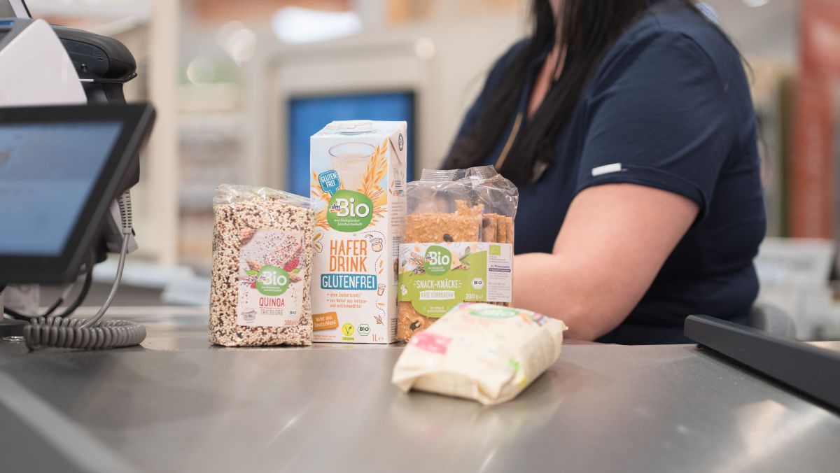 Drei Produkte auf einem Förderband an der Kassa - ein Haferdrink, eine Packung Quinoa sowie ein Snack-Knäckebrot.