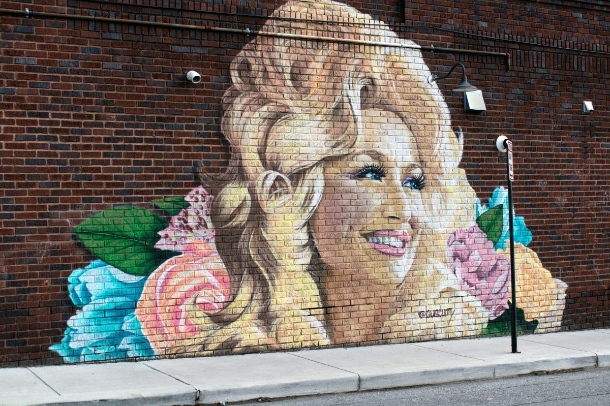 Eine Straße mit einer Backsteinmauer auf der ein Grafitti von Dolly Partons Gesicht, umgeben mit Blumen angebracht ist.