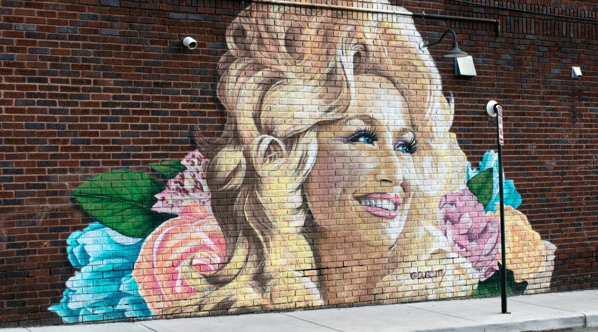 Eine Straße mit einer Backsteinmauer auf der ein Grafitti von Dolly Partons Gesicht, umgeben mit Blumen angebracht ist.